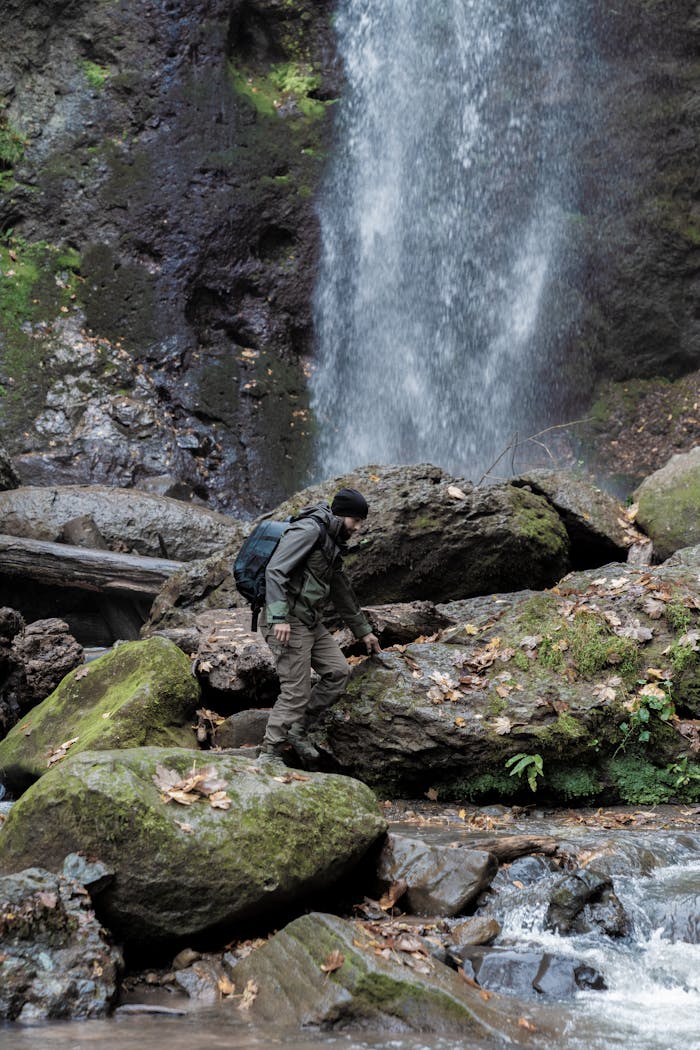 Adventurer climbs mossy rocks by a waterfall in a lush forest setting.
